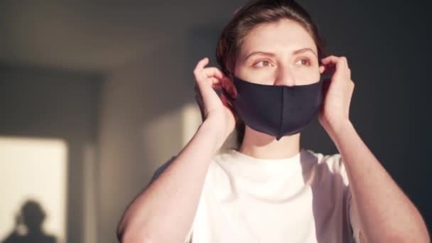 Eine junge Frau setzt zu Hause eine wiederverwendbare Schutzmaske auf und blickt in die Kamera. Coronavirus und Schutz vor Infektionen. Porträt eines maskierten Mädchens vor dem Gang nach draußen während der covid-19 — Stockvideo
