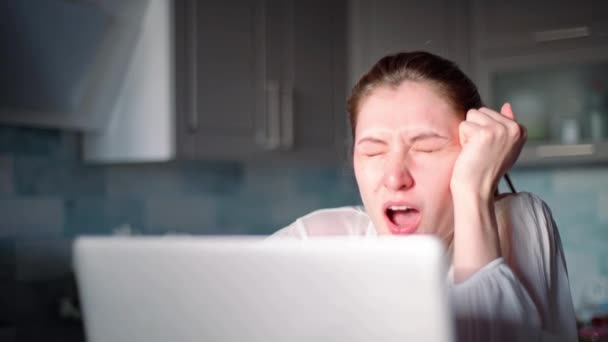 Freelancer boceja. Jovem mulher sentada na cozinha em casa parece cansada e bocejando enquanto trabalhava em um laptop. Negócios na internet, auto-isolamento e quarentena — Vídeo de Stock