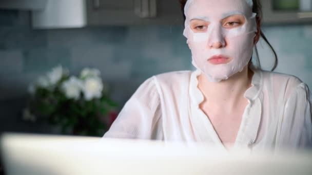 Женщина в косметической маске работает с ноутбуком дома на кухне — стоковое видео