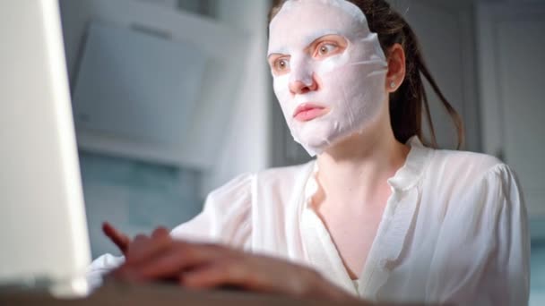 Cuidado facial de la piel en casa durante freelance. Mujer joven escribiendo en un teclado portátil con una máscara hidratante en la cara — Vídeo de stock