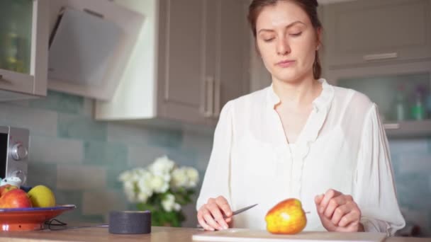 En ung kvinna lagar mat i sitt smarta kök med en bärbar högtalare och sitt röstrecept. Söt kock skär äpplen till ljudet av den personliga ljud assistent — Stockvideo