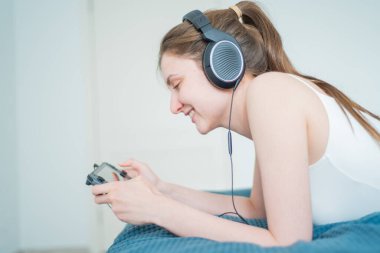 New York City, New York, ABD - 15 Mayıs 2020: Neşeli kız yatakta uzanıyor ve Sennheiser kulaklıklarıyla Nintendo Switch çalıyor. Dairede taşınabilir oyun