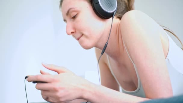 Una mujer con adicción al juego. Hermosa chica acostada juega en la consola con auriculares grandes — Vídeos de Stock