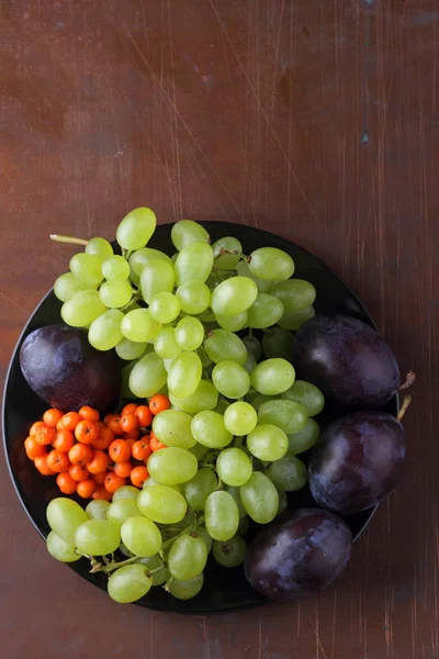 Grüne Trauben, blaue Pflaumen und orangefarbene Vogelbeeren auf einem schwarzen Teller für Designer im Retro-Stil — Stockfoto