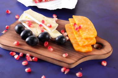 Camembert peyniri, peynir, turuncu peynir, mermer peynir, nar taneleri, koyu mavi üzüm ahşap oyun tahtası koyu mavi zemin üzerine dilimlenmiş