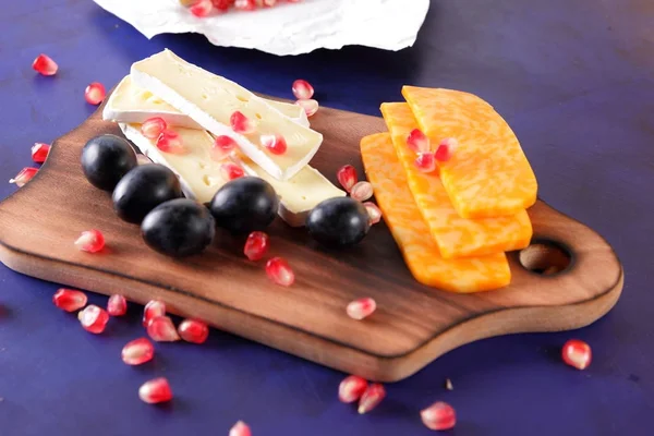 Camembert peyniri, peynir, turuncu peynir, mermer peynir, nar taneleri, koyu mavi üzüm ahşap oyun tahtası koyu mavi zemin üzerine dilimlenmiş