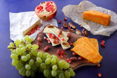 Camembert peyniri, turuncu sert peynir, Yeşil üzüm, fıstık, ceviz ve nar tohumları bir ahşap tahta üzerinde koyu mavi bir arka plan üzerinde