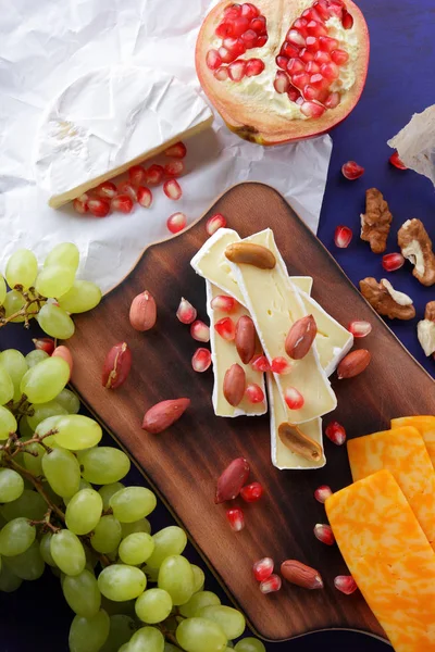 Camembert peyniri, turuncu sert peynir, Yeşil üzüm, fıstık, ceviz ve nar tohumları bir ahşap tahta üzerinde koyu mavi bir arka plan üzerinde