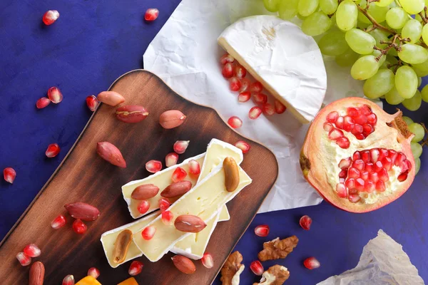 Camembert peyniri, turuncu sert peynir, Yeşil üzüm, fıstık, ceviz ve nar tohumları bir ahşap tahta üzerinde koyu mavi bir arka plan üzerinde