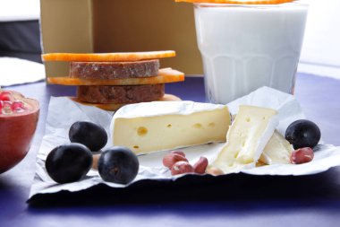 Camembert peyniri, turuncu mermer peynir, süt, nar, zeytin, fıstık tahıl, koyu mavi üzüm ve ekmek beyaz kumaş ve koyu mavi arka plan, peynir arka plan üzerine bir bardak