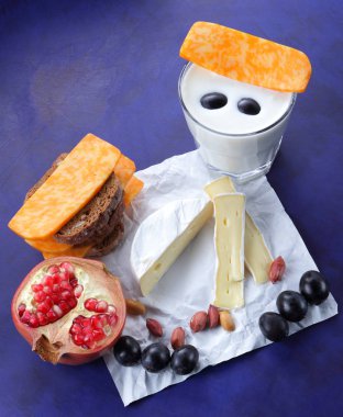 Camembert peyniri, turuncu mermer peynir, süt, nar, zeytin, fıstık tahıl, koyu mavi üzüm ve ekmek beyaz kumaş ve koyu mavi arka plan, peynir arka plan üzerine bir bardak