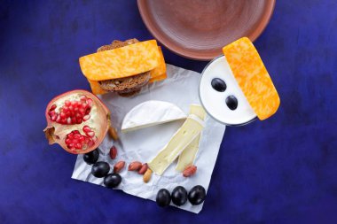 Camembert peyniri, turuncu mermer peynir, süt, nar, zeytin, fıstık tahıl, koyu mavi üzüm ve ekmek beyaz kumaş ve koyu mavi arka plan, peynir arka plan üzerine bir bardak