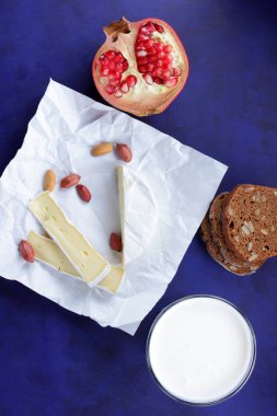 Camembert peyniri, bir bardak süt, nar, fıstık tahıl ve beyaz kumaş üzerine ekmek ve retro tarzı bir koyu mavi arka plan
