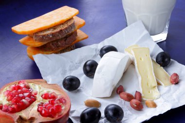 Camembert peyniri, turuncu mermer peynir, süt, nar, zeytin, fıstık tahıl, koyu mavi üzüm ve ekmek beyaz kumaş ve koyu mavi arka plan, peynir arka planda retro tarzı bir bardak