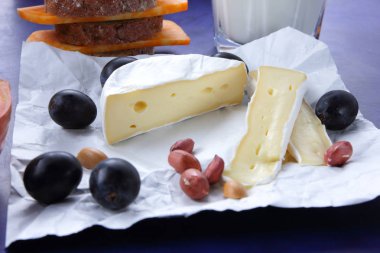 Camembert peyniri, turuncu mermer peynir, süt, nar, zeytin, fıstık tahıl, koyu mavi üzüm ve ekmek beyaz kumaş ve koyu mavi arka plan, peynir arka planda retro tarzı bir bardak