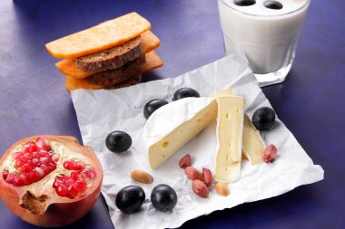 Camembert peyniri, turuncu mermer peynir, süt, nar, zeytin, fıstık tahıl, koyu mavi üzüm ve ekmek beyaz kumaş ve koyu mavi arka plan, peynir arka planda retro tarzı bir bardak