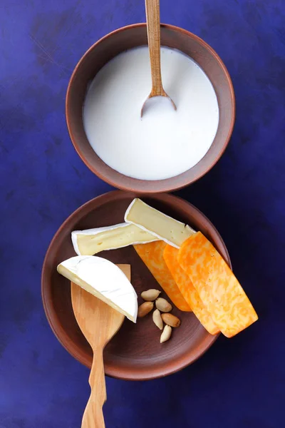 Queso camembert, queso naranja, cacahuetes, cuchara de madera y espátula con vajilla de barro sobre fondo azul oscuro —  Fotos de Stock