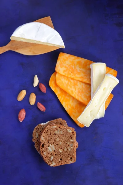 Camembert peyniri, turuncu peynir, fıstık, tahta spatula ve ekmek tahıl üzerinde koyu mavi arka plan ile