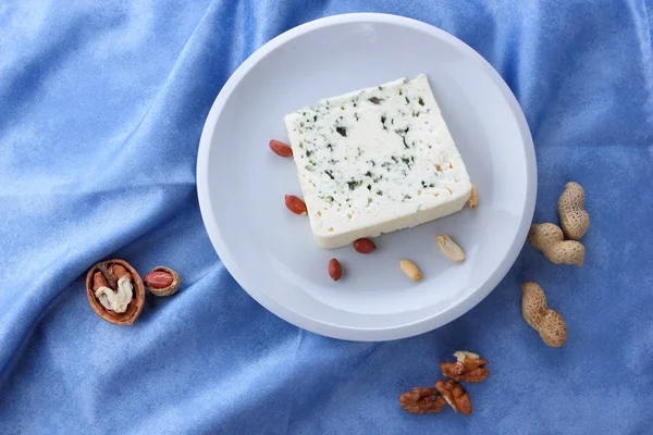 Formaggio con muffa e noci su un piatto bianco, formaggio di pecora su sfondo blu, formaggio e piatto bianco di arachidi su un tovagliolo blu, formaggio e noce in stile minimalista, stile retrò — Foto Stock