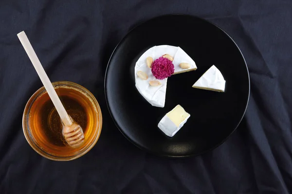 Cheese, camembert on a black plate, honey, pink flower, sliced cheese with mildew on a black velvet, minimalism, cheese on a black background