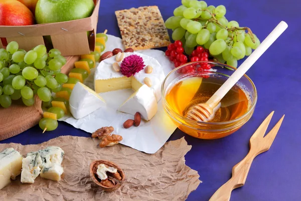 Camembert, cheese with mold, apples, green grapes, honey, red berries of viburnum, peanuts, dark blue background, pop art, retro style, pears, healthy food, cheddar on parchment paper, wooden board, art