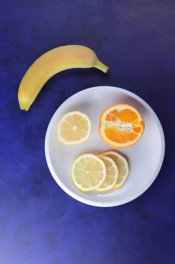 Ultraviyole arka planda minimalizm tarzı, dilim limon ve beyaz kaplama, sarı muz koyu mavi arka plan, sağlıklı gıda, vegan, pop sanat üzerinde yarım portakal narenciye