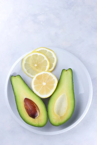 Avocado and slices of lemon in a white plate on a white background, green and yellow fruits, tropical fruits, sliced avocado and lemon on a marble background, healthy food, vegan, minimalism