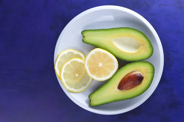 Avocado and slices of lemon on a white plate, tropical fruits on an ultraviolet background, vegetarian breakfast, Asian cuisine, avocado pop art, citrus with avocado