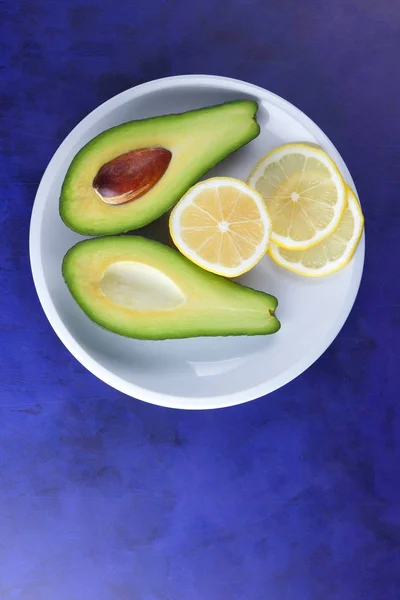 Avocado and slices of lemon on a white plate, tropical fruits on an ultraviolet background, vegetarian breakfast, Asian cuisine, avocado pop art, citrus with avocado