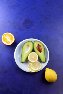 Avokado, limon ve beyaz bir plaka üzerinde yarım portakal, ultraviyole arka plan, vejetaryen Kahvaltı, Asya Mutfağı, avokado pop art, narenciye, turuncu avokado, sanat ile tropikal meyveler
