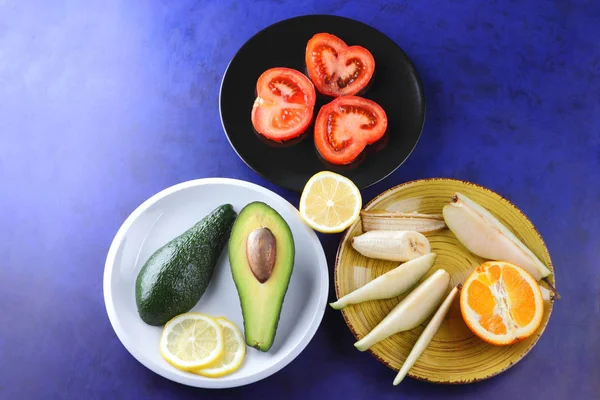 Avocado and lemon on a white plate, tomato on a black plate, citrus on an ultraviolet background, chopped banana and pear, avocado pop art, healthy food, vegan, retro style, pop art