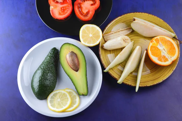 Avocado and lemon on a white plate, tomato on a black plate, citrus on an ultraviolet background, chopped banana and pear, avocado pop art, healthy food, vegan, pop art