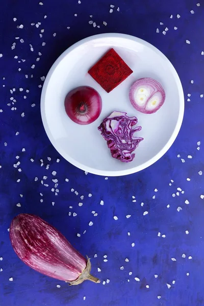 Purple cabbage, onions and beetroot on a white plate, vegetables in space, vegetables on the ultraviolet background, coarse salt, purple onions, purple eggplant on a dark blue background, pop art