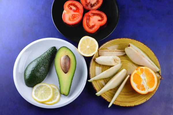 Avocado and lemon on a white plate, tomato on a black plate, citrus on an ultraviolet background, chopped banana and pear, avocado pop art, healthy food, vegan, art