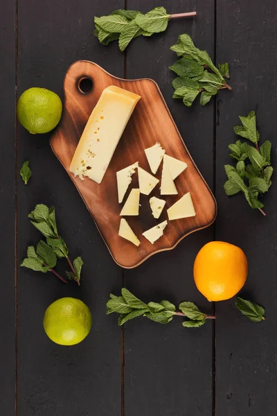 Hard yellow cheese, lemon, lime and fresh mint on a wooden black background, slices of cheese on a black background, top view, mint leaves with cheese with holes, rustic style, citrus, healthy food, vegan