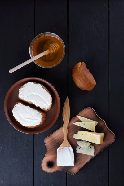 Hard cheese on a wooden board, sandwiches with soft cheese, honey in glass dishes, copy space, minimalism, cheese pattern, wooden dark background, healthy food, wooden cutlery
