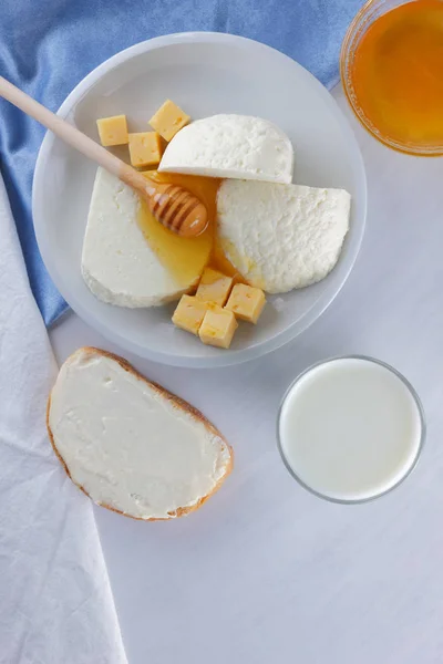 Cheese, milk assortment on white background, white cheese and honey on a blue napkin, wooden background, wooden stick for honey, top view, cheese on white plate, kefir in a glass