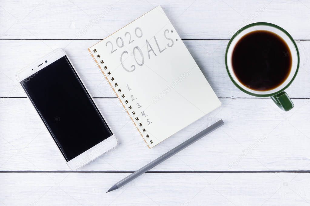 2020 new year goals, plan, action text on notepad on white wooden boards. 2020 goals on blank note paper with copy space for text and smartphone. Cup of coffee over wooden boards