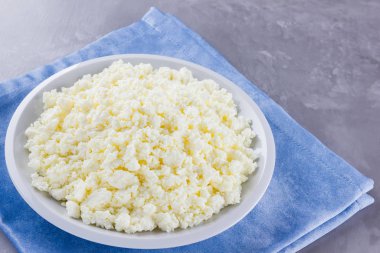 Tabakta süzme peynir. Gri arka planda beyaz tabakta taze süzme peynir. Mavi peçetede yumuşak peynir. Üst Manzara. Boşluğu kopyala