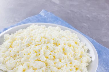 Tabakta süzme peynir. Gri arka planda beyaz tabakta taze süzme peynir. Mavi peçetede yumuşak peynir. Üst Manzara. Boşluğu kopyala