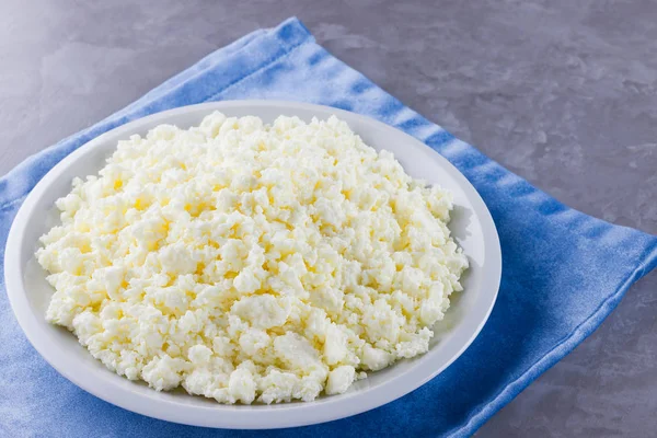 Cottage cheese in a plate. Fresh cottage cheese in white plate on a gray background. Soft cheese on a blue napkin. Top view. Copy space