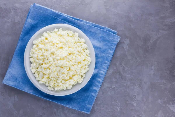 Cottage cheese in a plate. Fresh cottage cheese in white plate on a gray background. Soft cheese on a blue napkin. Top view. Copy space