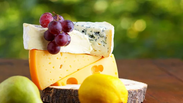 Camembert, brie, hard cheese and grapes on wooden stand. Pieces of different cheeses, pear and lemon on a blurred background. Dorblu and soft cheese on wooden boards. Widescreen
