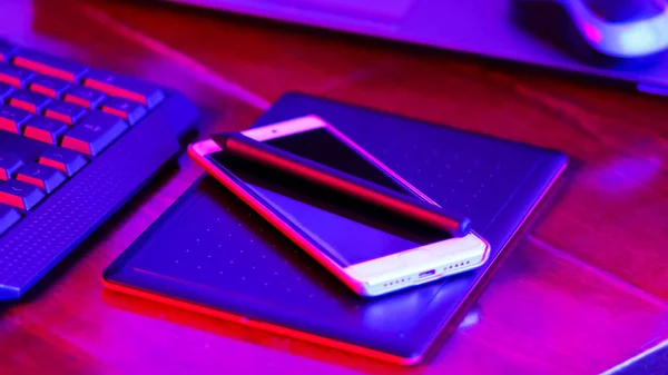 Freelancer workplace in neon light. Computer, graphics tablet and smartphone on the table. Place for creativity in blue pink light. Widescreen