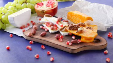 Peynir tabağı: ahşap tabakta meyve ve fındıklı peynir çeşitleri. Farklı peynir türleri. Klasik mavi arka planda Camembert, üzüm ve nar. Üst görünüm