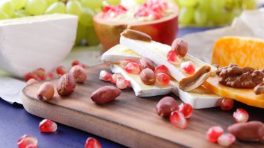 Peynir tabağı: ahşap tabakta meyve ve fındıklı peynir çeşitleri. Farklı peynir türleri. Klasik mavi arka planda Camembert, üzüm ve nar.