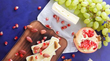 Peynir tabağı: ahşap tabakta meyve ve fındıklı peynir çeşitleri. Farklı peynir türleri. Klasik mavi arka planda Camembert, üzüm ve nar.