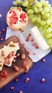 Peynir tabağı: ahşap tabakta meyve ve fındıklı peynir çeşitleri. Farklı peynir türleri. Klasik mavi arka planda Camembert, üzüm ve nar.