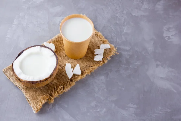 Coconut milk and coconut in paper glass on gray background. Coconut vegan milk non dairy on sackcloth. Healthy drink concept. Alternative milk. Top view