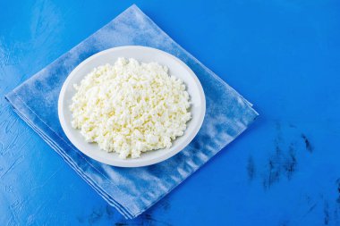 Mavi arka planda beyaz tabakta süzme peynir. Organik ekolojik sağlıklı yemek, süt ürünleri. Mavi peçeteye bir tabak süzme peynir. Üst Manzara. Boşluğu kopyala
